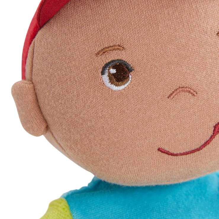 Nahaufnahme eines Plüschpuppengesichts mit braunen gestickten Augen, einer kleinen Nase, einem lächelnden Mund, einem roten Hut und einem blauen Hemd mit gelben Ärmeln. Nur ein Teil des Gesichts und des Oberkörpers sind sichtbar.