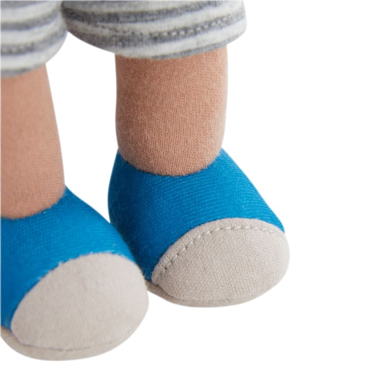 Nahaufnahme eines Puppenbeins, das hellbraune Strümpfe, blaue Schuhe mit beigen Zehen und eine grau-weiß gestreifte Hose trägt. Der Hintergrund ist weiß.
