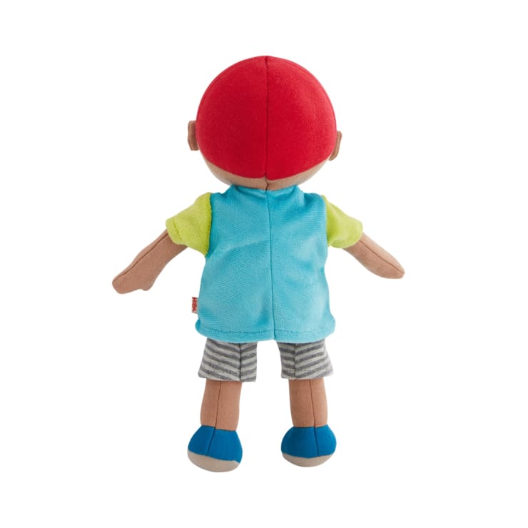 Eine ausgestopfte Puppe wird von hinten gezeigt, die eine rote Mütze, eine blaue Weste, ein gelbes Hemd, eine grau gestreifte kurze Hose und blaue Schuhe trägt. Die Puppe steht aufrecht mit leicht ausgestreckten Armen.