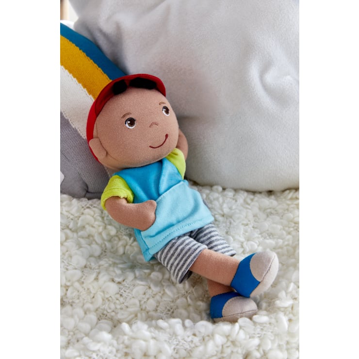 Eine Plüschpuppe mit brauner Haut, die eine rote Mütze, eine blaue Weste, ein grünes Hemd, gestreifte Shorts und blaue Schuhe trägt, liegt auf einer strukturierten weißen Decke mit einem weißen Kissen und einem gestreiften Kissen dahinter.