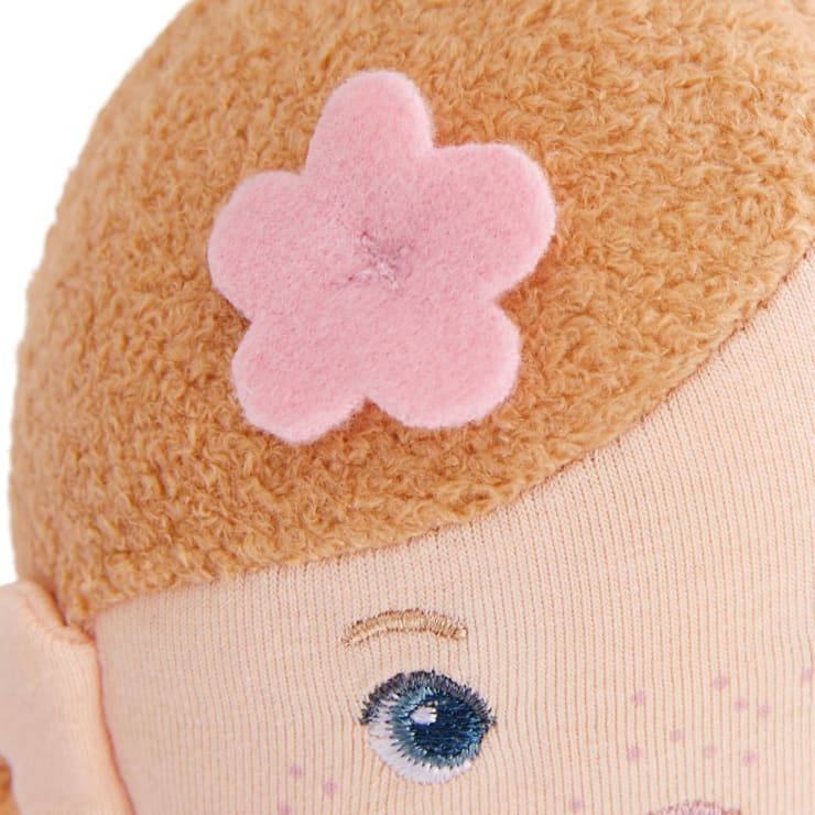 Nahaufnahme eines Puppenkopfes aus weichem Stoff mit einer rosafarbenen Filzblume, die an lockigem hellbraunem Haar befestigt ist. Ein Teil der gestickten blauen Augen und Ohren ist sichtbar.