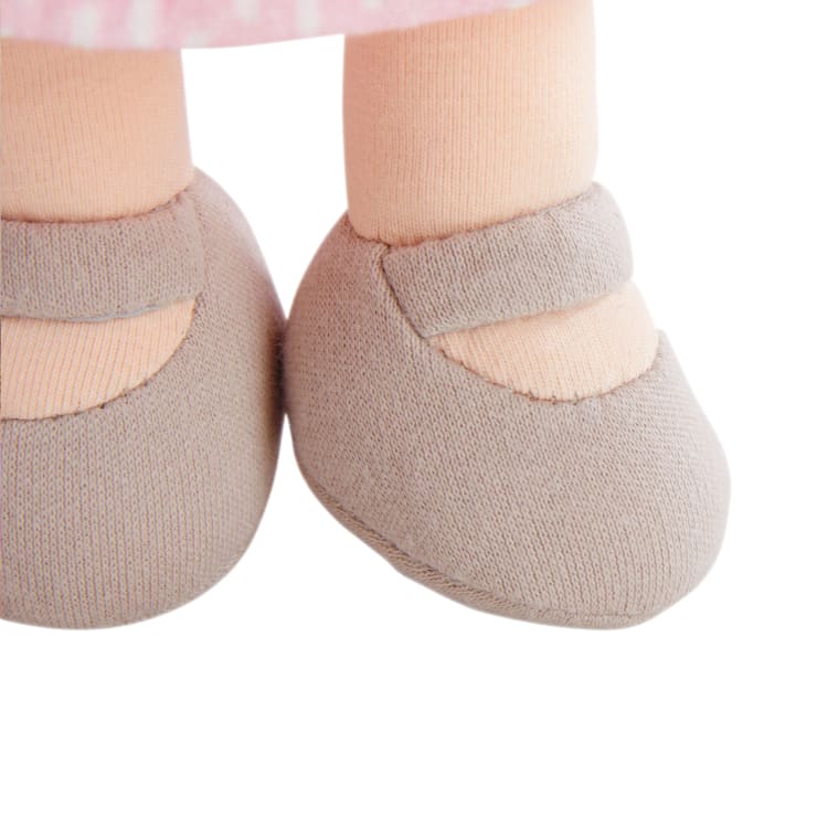 Nahaufnahme eines Puppenfußes, der hellbraune Stoffschuhe mit Riemen und pfirsichfarbene Socken trägt, die teilweise von einem rosa gemusterten Rock bedeckt sind.