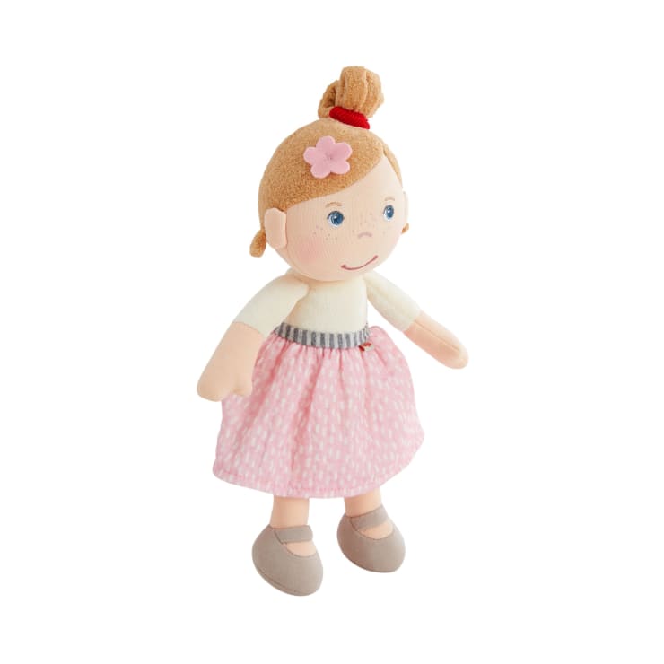 Plüschpuppe mit blondem Haar in einem Dutt, rosa Blumenhaarspange, weißem Oberteil, rosa gemustertem Rock und beigen Schuhen, die aufrecht vor einem weißen Hintergrund steht.