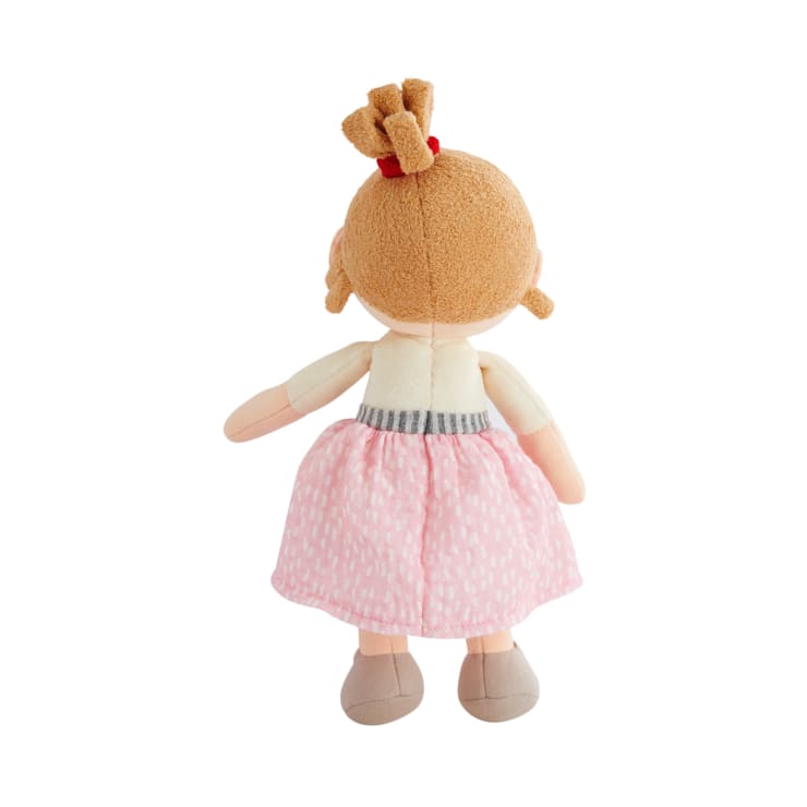 Eine Plüschpuppe mit Blickrichtung nach außen, die einen rosa gemusterten Rock, beigefarbene Schuhe und ein cremefarbenes Oberteil trägt. Die Puppe hat hellbraunes Haar, das zu einem Dutt mit einem roten Band frisiert ist. Der Hintergrund ist einfarbig weiß.