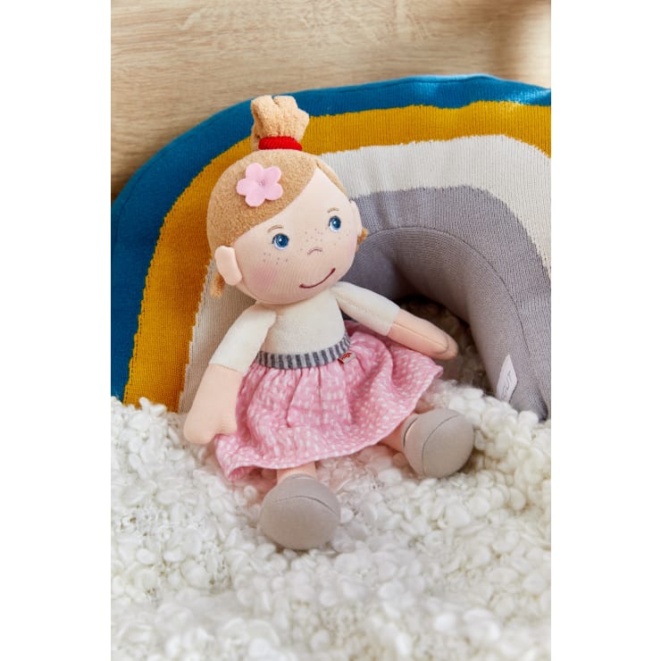 Eine weiche Stoffpuppe mit blauen Augen, einem rosa Rock und einem Blumenhaarschmuck sitzt auf einer weißen, strukturierten Decke. Hinter der Puppe befindet sich ein mehrfarbiges Kissen mit blauen, gelben und grauen Streifen.