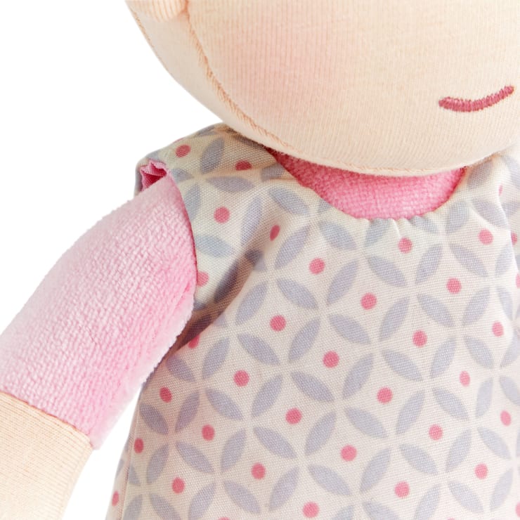 Nahaufnahme einer Stoffpuppe, die ein gemustertes Kleid mit kreisförmigen Mustern in Grau und Rosa und darunter ein rosa Hemd trägt. Das Bild zeigt die Schulter, den Arm, den Hals und einen Teil des lächelnden Gesichts der Puppe.