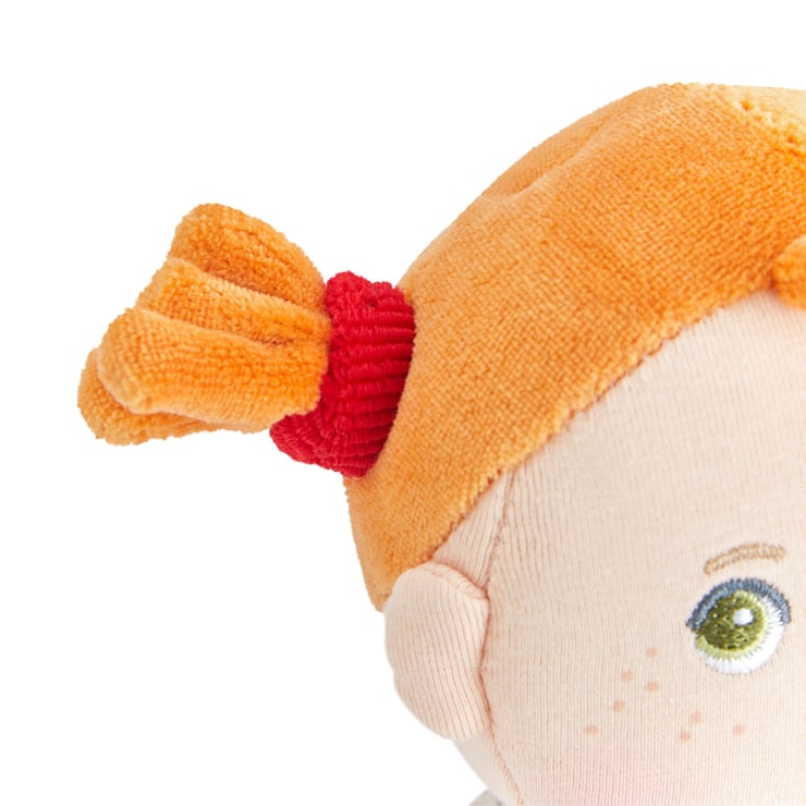 Nahaufnahme eines Stoffpuppenkopfes mit orangefarbenem Haar, das mit einem roten Haarband zu einem Pferdeschwanz gebunden ist. Die Puppe hat gestickte Gesichtszüge, darunter ein grünes Auge, eine Augenbraue und Sommersprossen auf hellem Stoff.