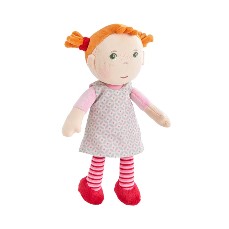 Eine Stoffpuppe mit orangefarbenen Haaren in Zöpfen, grünen Augen und Sommersprossen, die ein grau gemustertes Kleid, ein rosa Hemd, rot-weiß gestreifte Leggings und rote Schuhe trägt, steht vor einem weißen Hintergrund.