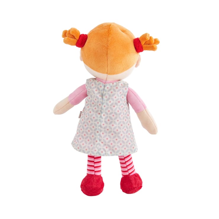 Eine Plüschpuppe von hinten gesehen, mit orangefarbenen Haaren in Zöpfen, einem gemusterten Kleid, rosa Ärmeln, rot-weiß gestreiften Leggings und roten Schuhen, vor weißem Hintergrund.
