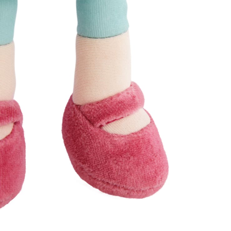 Nahaufnahme der Unterschenkel einer Puppe, die hellblaue Leggings und beigefarbene Socken trägt, mit rosa Plüschschuhen, die oben einen Riemen haben, vor einem weißen Hintergrund.