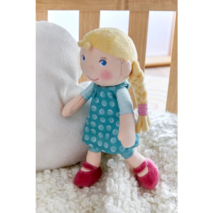 Eine weiche Stoffpuppe mit blondem geflochtenem Haar, die ein blaues Kleid mit weißen Punkten, ein hellblaues Hemd und rote Schuhe trägt, sitzt auf einer weißen strukturierten Decke neben einem weißen Kissen.