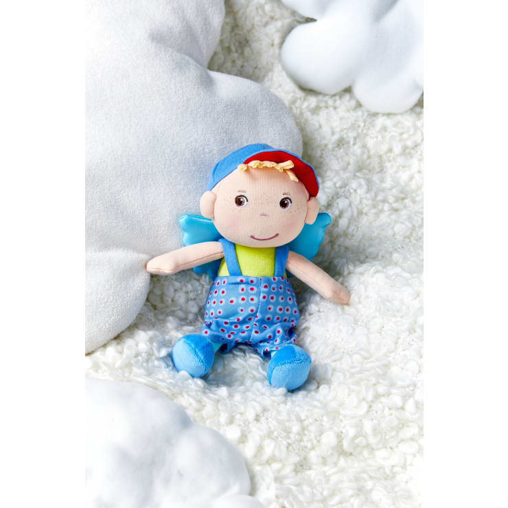 Eine weiche Stoffpuppe mit blauen Flügeln, einem blauen Overall mit roten und weißen Punkten, einer blau-roten Mütze und einem gelben Hemd, die auf einer weißen, strukturierten Oberfläche mit weißen Plüschwolken in der Nähe liegt.