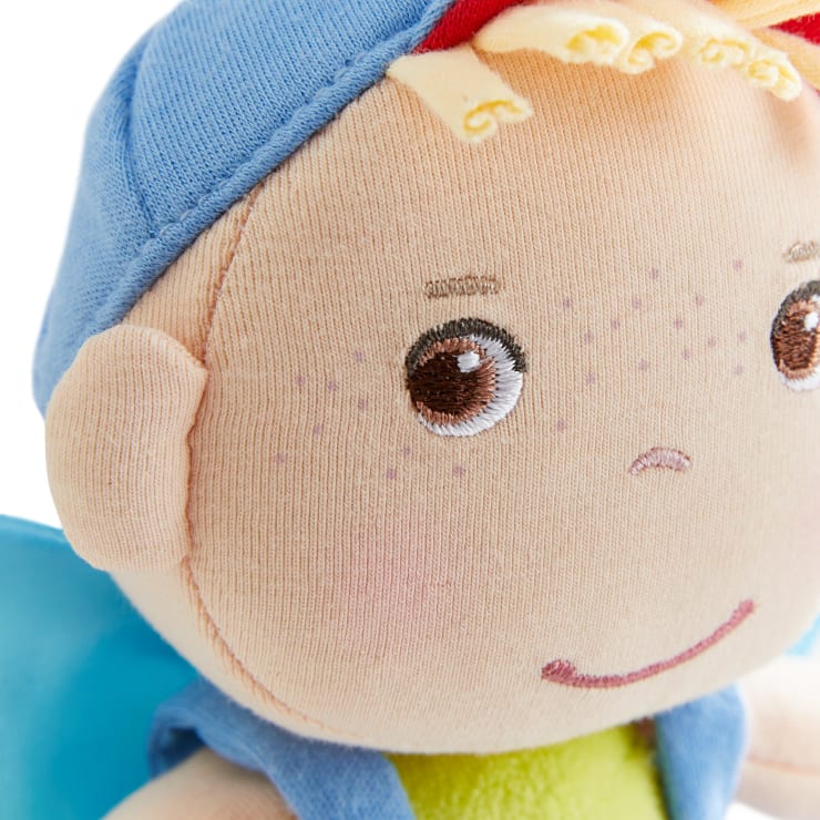 Nahaufnahme einer weichen Stoffpuppe mit braunen gestickten Augen, einem kleinen gestickten Lächeln und hellbraunen gepunkteten Sommersprossen. Die Puppe trägt einen blauen Hut, hat hellgelbe Garnhaare und ein blaues Hemd.