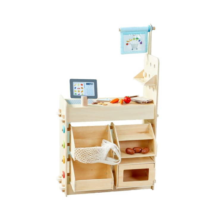Ein hölzernes Spielset für einen Lebensmittelladen mit einer Registrierkasse, einer blauen Flagge, Behältern mit einem Netzbeutel, Spielzeuggemüse und Regalen zur Aufbewahrung, alles vor einem weißen Hintergrund.