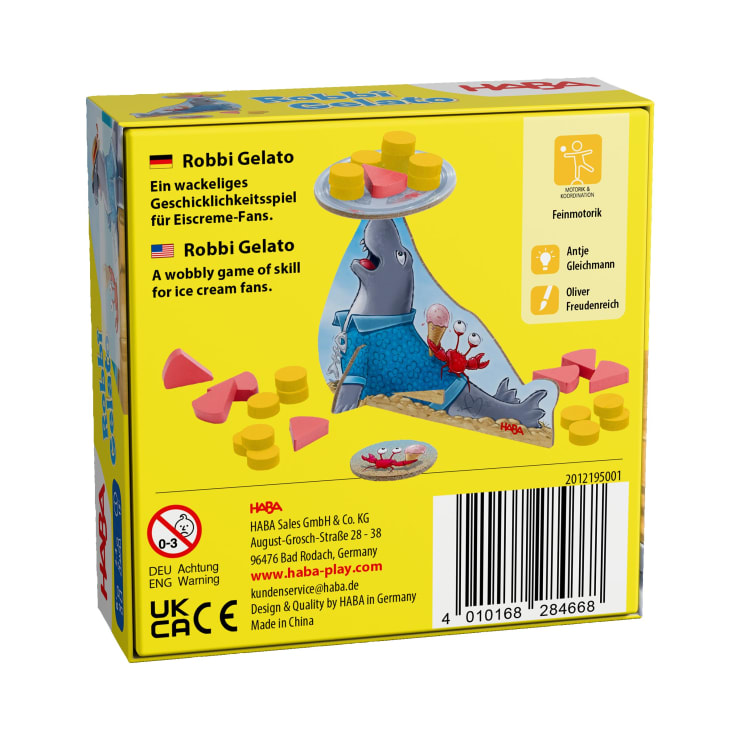 Eine gelbe Schachtel für das Spiel Robbi Gelato von HABA zeigt eine graue Robbe, die eine Eisschale auf der Nase balanciert und von gelben und rosa Spielsteinen umgeben ist. Auf der Schachtel sind eine Anleitung und Informationen zum Unternehmen aufgedruckt.