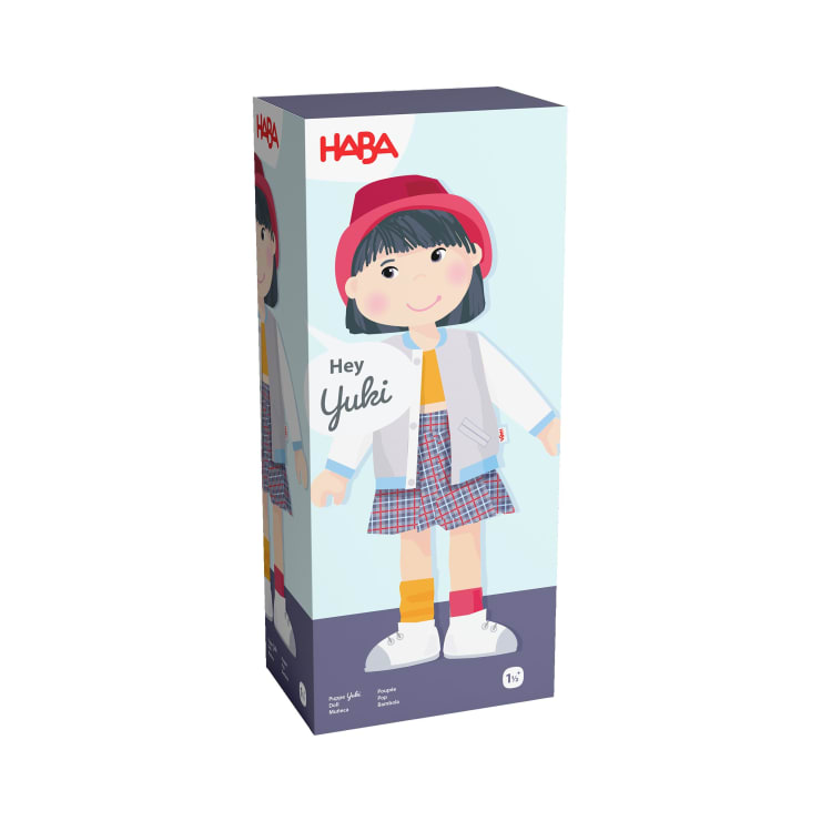 Eine rechteckige Schachtel mit einer illustrierten Puppe mit schwarzen Haaren, einem roten Hut, einer hellblauen Jacke, einem karierten Rock und Turnschuhen. Der Text auf der Schachtel lautet: HABA und Hey Yuki. Geeignet für Kinder ab 1,5 Jahren.