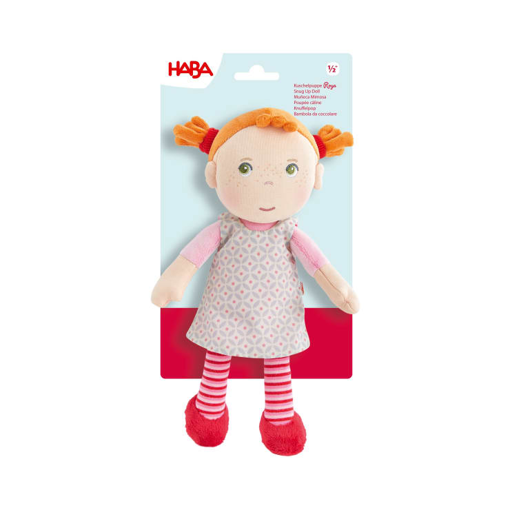 Eine weiche Stoffpuppe mit orangefarbenen Zöpfen, die ein gemustertes graues Kleid, ein rosa Hemd, gestreifte Leggings und rote Schuhe trägt, wird vor einer Verpackung mit dem HABA-Logo präsentiert.