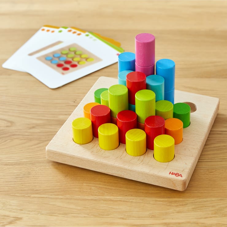 Ein hölzernes Stapelspielzeug mit farbigen zylindrischen Teilen, die in Reihen auf einer quadratischen Grundfläche angeordnet sind. Im Hintergrund sind mehrere Aufgabenkarten mit farbigen Kreisen auf einer Holzfläche zu sehen.