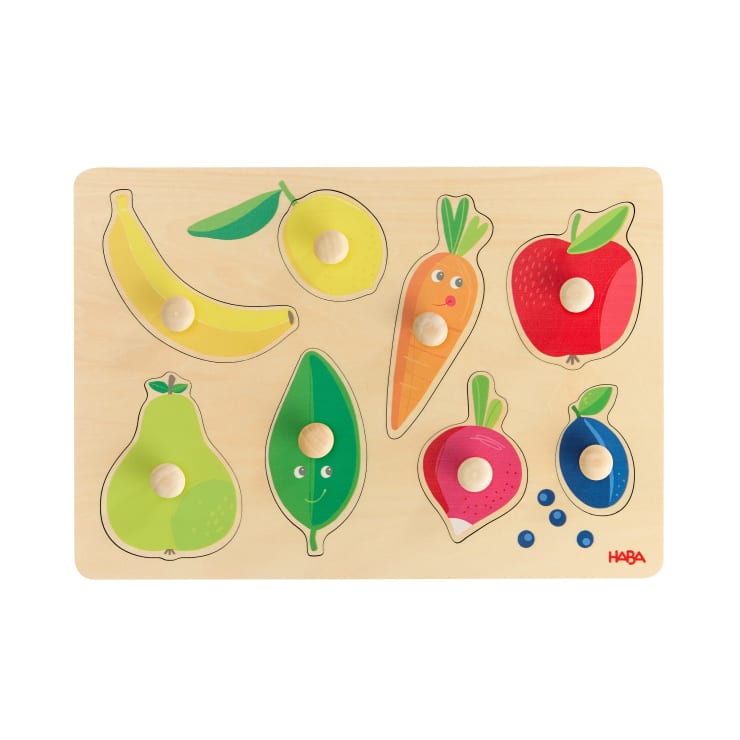 Ein hölzernes Puzzlebrett mit genoppten Teilen in Form einer Banane, einer Zitrone, eines Apfels, einer Birne, einer Erbsenschote, einer Karotte, einer Erdbeere und einer Blaubeere, die jeweils in die entsprechenden Ausschnitte passen.