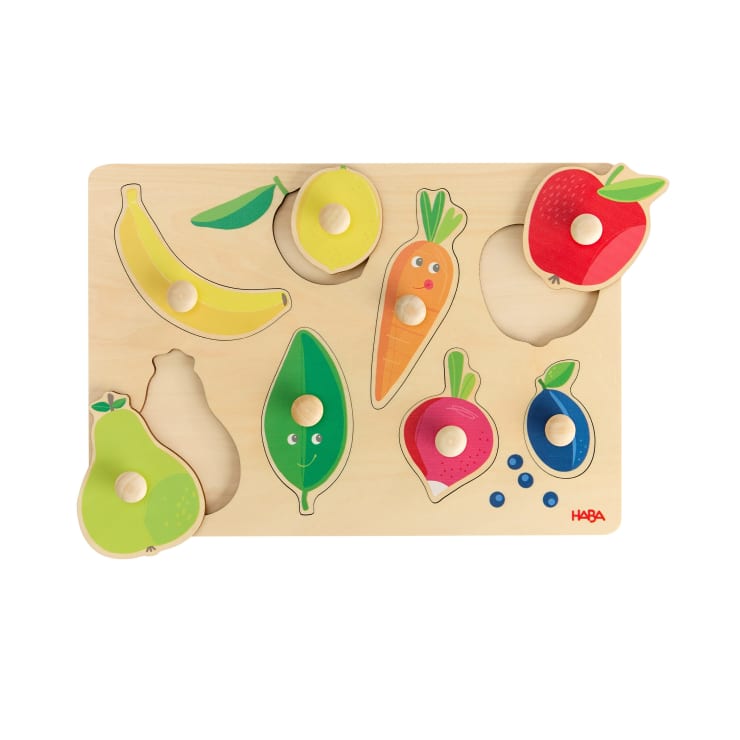 Ein hölzernes Puzzlebrett mit herausnehmbaren Teilen in Form einer Banane, eines Apfels, einer Birne, einer Karotte, eines Blattes, einer Erbsenschote, eines Rettichs und einer Blaubeere, jeweils mit einem Knopf zur einfachen Handhabung. Einige Teile haben einfache Gesichter.