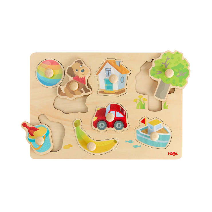 Ein hölzernes Puzzlebrett mit sechs Teilen: ein Ball, ein Hund, ein Haus, ein Baum, ein Spielzeugeimer mit Schaufel, ein rotes Auto und eine Banane. Jedes Teil hat einen Knauf zum einfachen Greifen. Das Brett hat passende Ausschnitte.