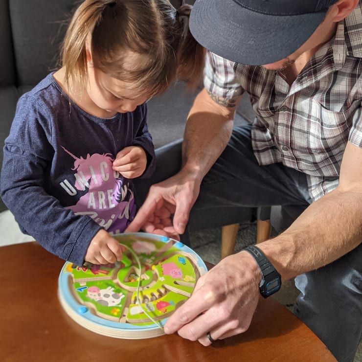 Ein junges Mädchen und ein Erwachsener sitzen an einem Tisch und bewegen mit ihren Händen magnetische Stäbe über ein kreisförmiges Bauernhof-Labyrinthspiel. Das Sonnenlicht scheint auf die Szene. Der Erwachsene trägt eine Mütze und eine Uhr.