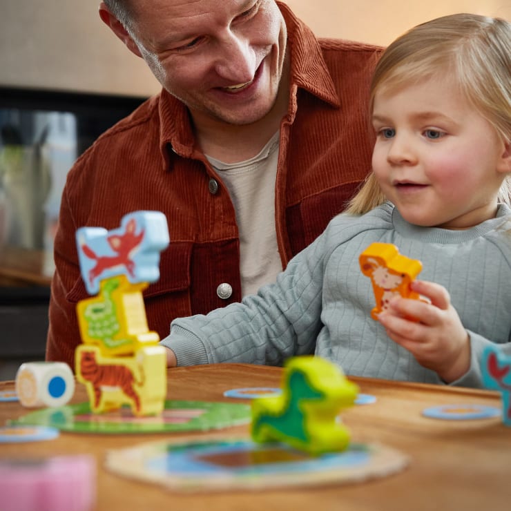 Ein Erwachsener und ein kleines Kind sitzen an einem Tisch und spielen ein Brettspiel mit bunten Tierfiguren. Der Erwachsene lächelt, während das Kind einen Spielstein in Form eines Tieres in der Hand hält und andere Spielsteine auf dem Tisch verstreut sind.