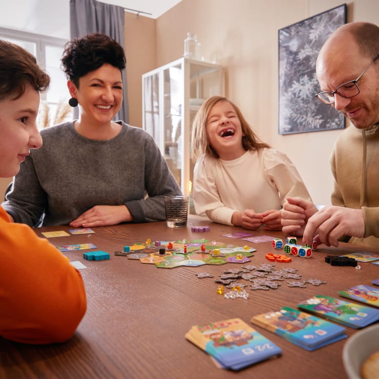 Vier Personen sitzen an einem Tisch und spielen ein Brettspiel. Spielkarten, Würfel und Spielfiguren sind auf dem Tisch verteilt. Ein Mädchen lacht, und die anderen lächeln oder sind konzentriert. Die Umgebung scheint ein helles, gemütliches Haus zu sein.