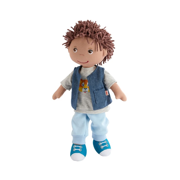 Eine weiche Stoffpuppe mit braunem Garnhaar, die ein graues T-Shirt mit einer Hundegrafik, eine blaue Weste, eine hellblaue Hose und blaue Turnschuhe trägt und aufrecht vor einem weißen Hintergrund steht.