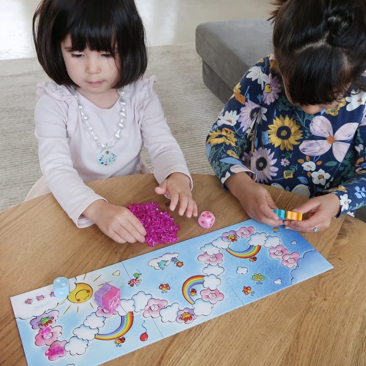 Zwei kleine Kinder sitzen an einem Holztisch und spielen ein farbenfrohes Brettspiel mit Abbildungen von Regenbögen, Wolken und der Sonne. Ein Kind hat rosa Kristalle in der Hand, während das andere orangefarbene und gelbe Steine hält.