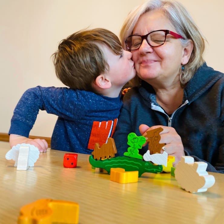 Ein kleiner Junge küsst eine ältere Frau auf die Wange, während sie an einem Tisch sitzen und ein Brettspiel mit hölzernen Tierfiguren spielen. Beide lächeln. Die Frau trägt eine Brille und einen Kapuzenpulli. Die Szene wirkt fröhlich und warm.