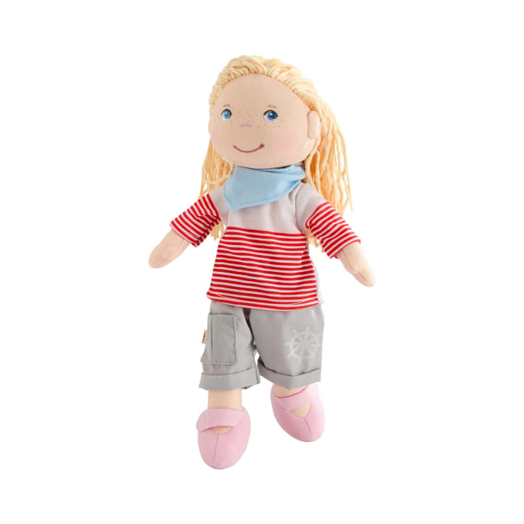 Eine weiche Stoffpuppe mit heller Haut und blondem Garnhaar, die ein rot-weiß gestreiftes Hemd, einen hellblauen Schal, eine graue Hose mit weißem Muster und rosafarbene Schuhe trägt, dargestellt vor einem weißen Hintergrund.