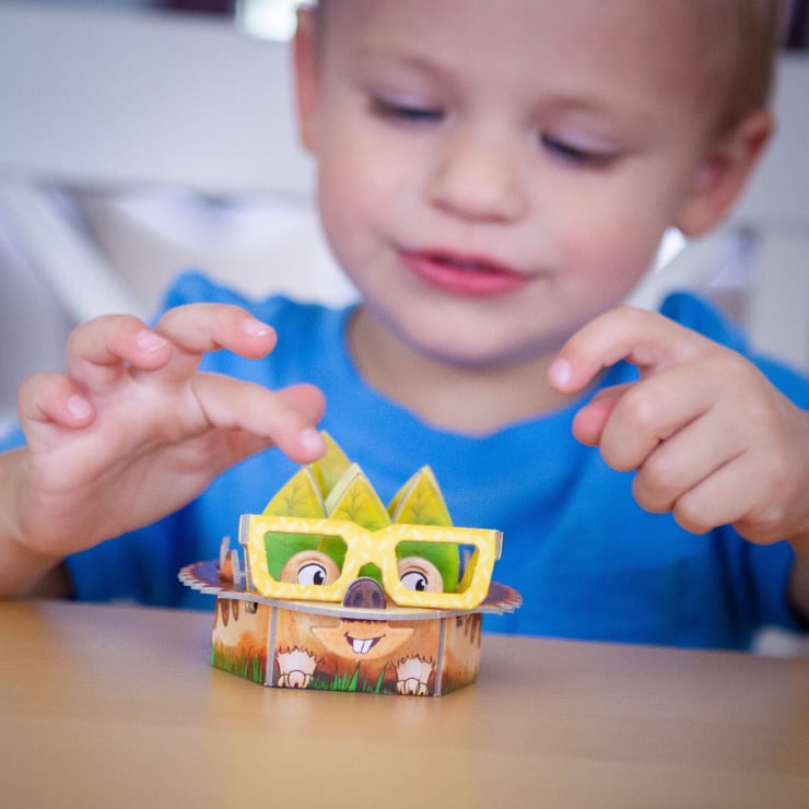 Ein kleines Kind in einem blauen Hemd sitzt an einem Tisch und betrachtet und spielt mit einer gelben Brille mit einem kleinen Tiermotiv aus Papier.