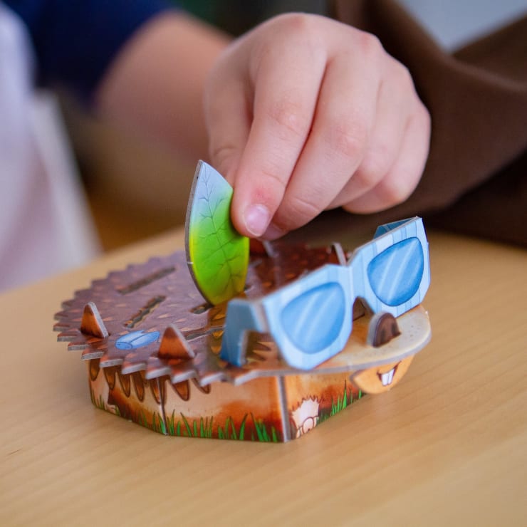 Eine Kinderhand legt ein grünes, blattförmiges Teil auf ein igelförmiges Brettspiel oder Spielzeug mit blauer Sonnenbrille und braunem, stacheligem Rücken, das auf einem Holztisch liegt.