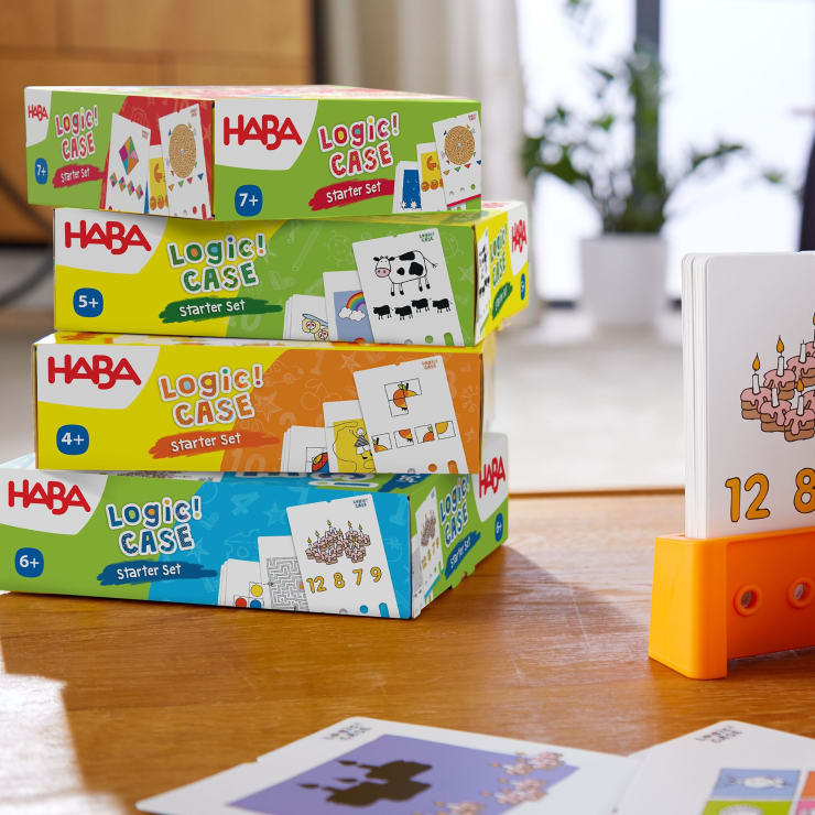 Drei HABA Logic! CASE Starter Set sind auf einem Tisch gestapelt, jede mit einer anderen Farbe und Altersbeschriftung. Im Vordergrund steht ein Ständer mit einer Karte, auf der eine Torte mit Kerzen und den Zahlen 12 und 8 abgebildet ist.