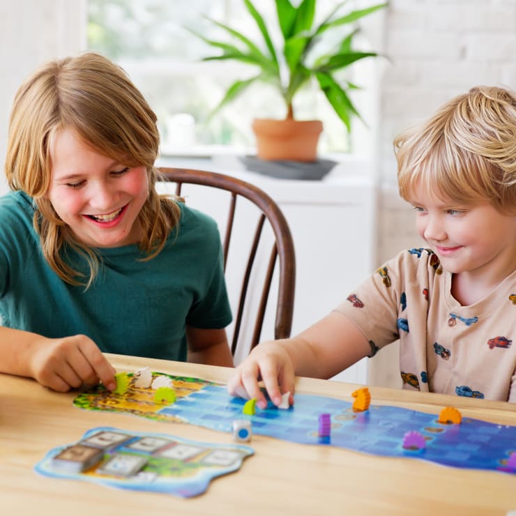 Zwei Kinder sitzen an einem Holztisch, lächeln und spielen zusammen ein buntes Brettspiel. Im Hintergrund sind eine Topfpflanze und ein Fenster zu sehen, die eine helle, freundliche Innenumgebung schaffen.