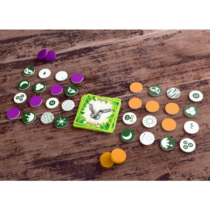 Eine Karte mit einer Eule ist von runden Spielsteinen mit verschiedenen Symbolen umgeben. Die Spielsteine sind in Gruppen mit grünen, orangen, lila und weißen Farben auf einer Holzoberfläche angeordnet.