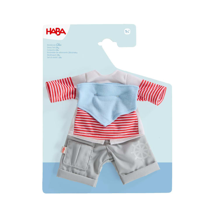 Ein verpacktes Puppen-Outfit-Set von HABA mit einem rot-weiß gestreiften langärmeligen Hemd, einem hellblauen Halstuch und einer hellgrauen Hose mit weißem Schiffsrad-Motiv auf blau-weißem Hintergrund.