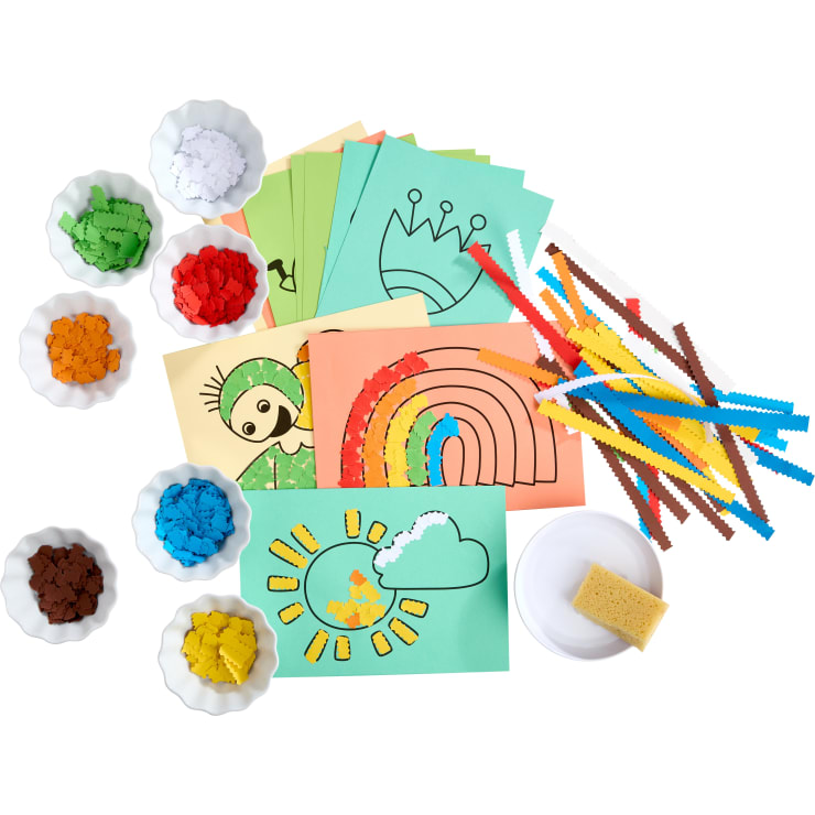 Buntes Bastelpapier mit skizzierten Mustern, Schalen mit farbigen Papierstücken, gewellte Papierstreifen, ein kleiner Schwamm und eine weiße Schale werden auf einer weißen Fläche für eine Bastelaktivität für Kinder angeordnet.