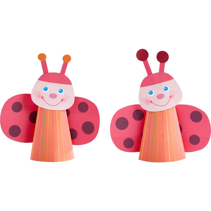 Zwei Marienkäfer aus Papier mit rosa Flügeln und roten Flecken, lächelnden Gesichtern, blauen Augen und gestreiften Kegelkörpern. Jeder hat zwei Fühler mit runden Spitzen und runde, errötende Wangen.