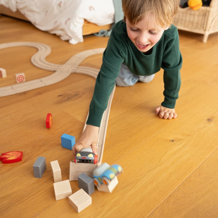 Ein kleines Kind in einem grünen Pullover spielt auf einem Holzboden und schiebt eine Spielzeugeisenbahn von einer Holzschiene in einen Stapel von Holzklötzen. Im Hintergrund sind weitere Spielzeuge und ein Bett zu sehen.