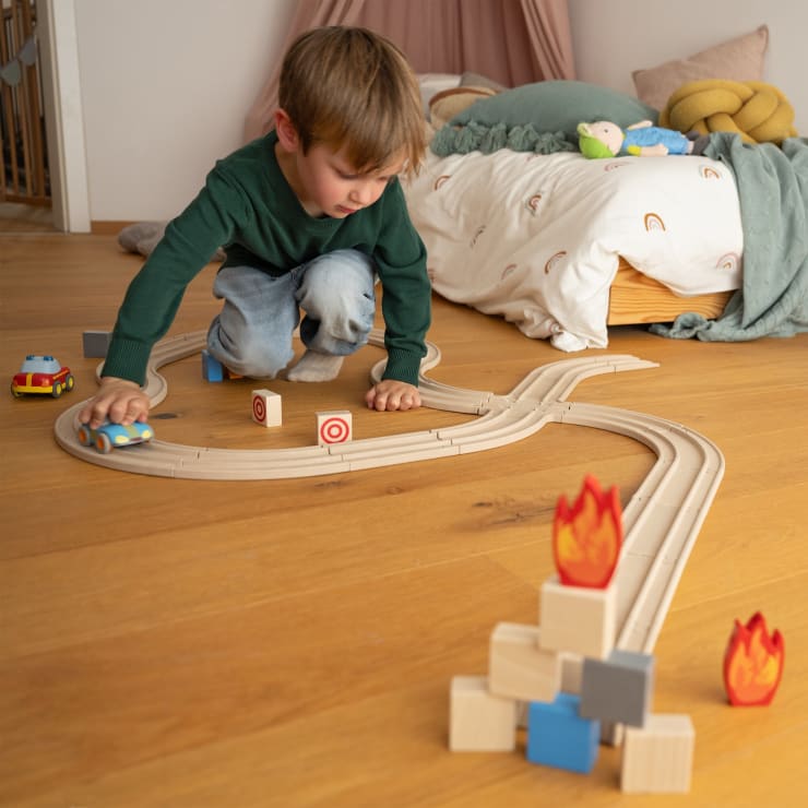 Ein kleines Kind in grünem Hemd und Jeans spielt mit einer hölzernen Spielzeugautobahn auf einem Holzboden neben einem Bett, auf der Spielzeugflammen und Zielblöcke angeordnet sind.