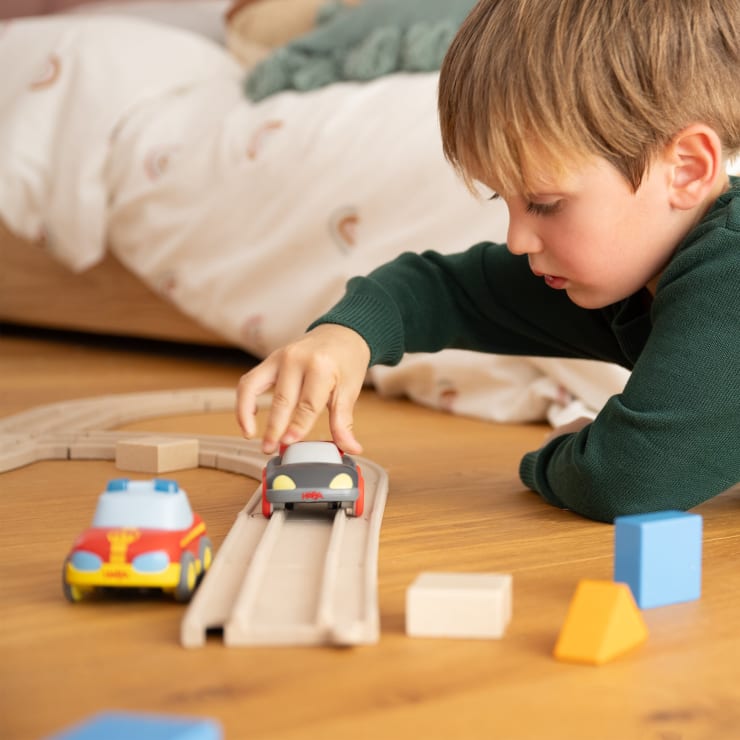 Ein kleiner Junge in einem grünen Hemd liegt auf dem Boden und spielt mit Spielzeugautos auf einer Holzbahn. Um ihn herum sind verschiedene Holzklötze verstreut, und im Hintergrund ist ein Bett zu sehen.