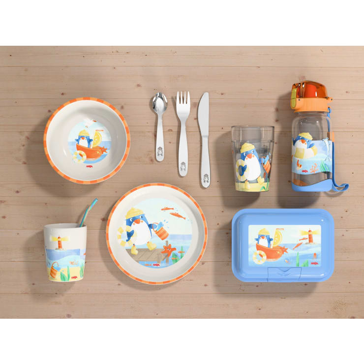 Kindergeschirr mit Pinguin-Motiv, bestehend aus Tellern, einer Schüssel, Trinkbechern, Utensilien, einer Brotdose und einer Wasserflasche, alles auf einer hellen Holzoberfläche.