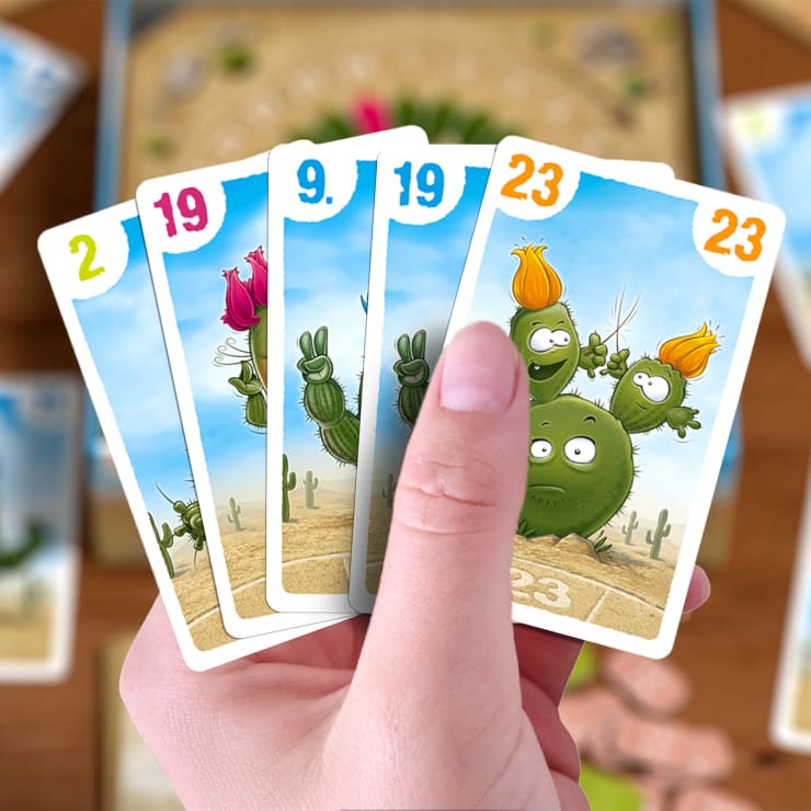 Eine Hand hält fünf bunte Spielkarten, auf denen Cartoon-Kakteen mit Zahlen abgebildet sind: 2, 19, 9, 19 und 23. Der Hintergrund zeigt ein verschwommenes Brettspiel mit weiteren Karten und Figuren.