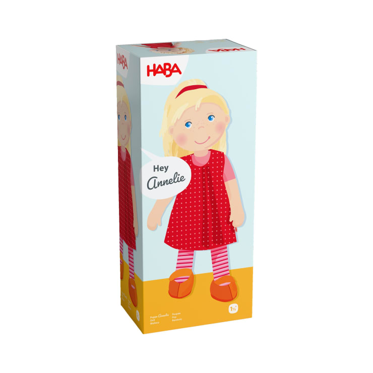 Eine rechteckige Schachtel mit dem HABA-Logo an der Oberseite zeigt die Illustration einer blonden Puppe in einem roten Kleid, einer gestreiften Strumpfhose und roten Schuhen. Die Schachtel trägt die Aufschrift "Hey Annelie" und ist für Kinder ab 1½ Jahren gedacht.