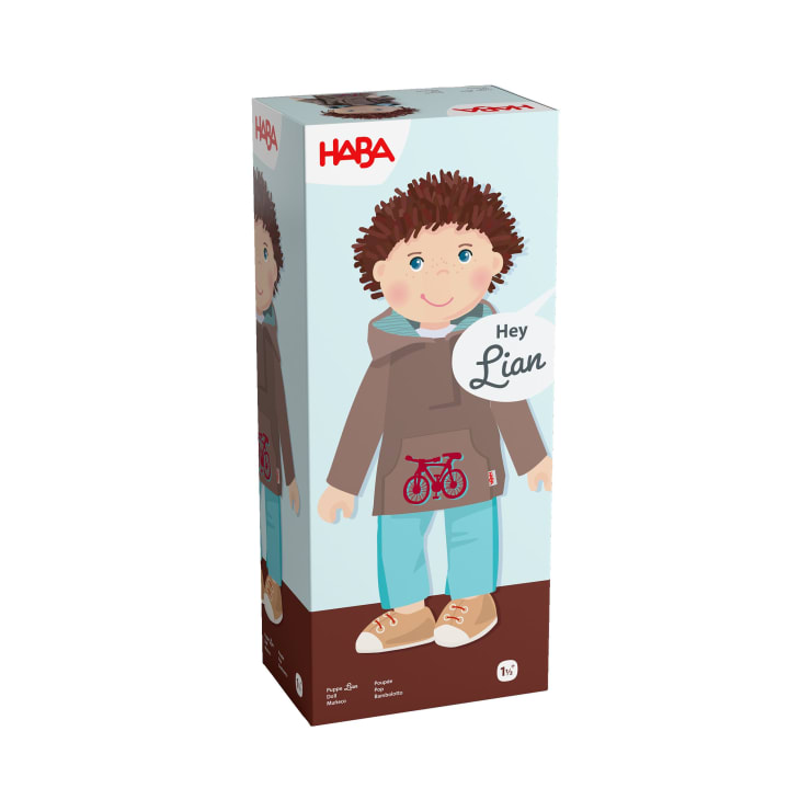 Eine rechteckige Schachtel zeigt eine Puppe namens Lian von HABA. Die Schachtel zeigt eine illustrierte Puppe mit braunen Haaren, blauen Augen, einem braunen Kapuzenpulli mit roter Fahrradgrafik, einer blauen Hose und braunen Schuhen.