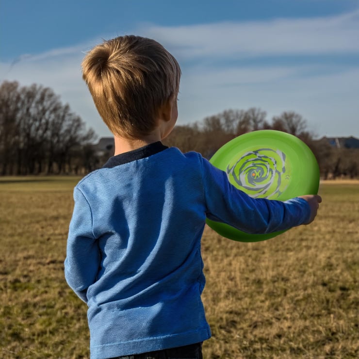 Ein kleines Kind mit hellbraunem Haar, das ein blaues langärmeliges Hemd trägt, steht auf einem großen offenen Feld und hält einen grünen Frisbee. Im Hintergrund sind Bäume und Gebäude unter einem teilweise bewölkten Himmel zu sehen.