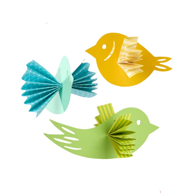 Drei Papiervögel aus farbigem und gemustertem Papier sind auf einem weißen Hintergrund angeordnet. Die Vögel sind gelb, grün und blau, jeweils mit akkordeonartig gefalteten Flügeln.
