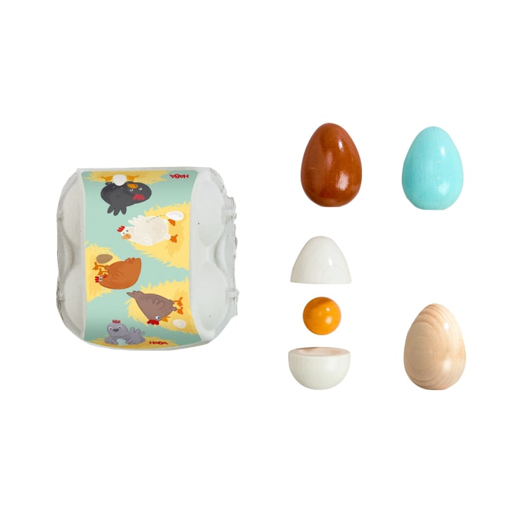 Ein Spielzeugset mit einem weißen Eierkarton mit einem Cartoon-Hühnermotiv und vier Holzeiern - braun, blau, weiß und aus Naturholz. Das weiße Ei ist aufgeschlagen und zeigt ein gelbes Eigelb im Inneren.